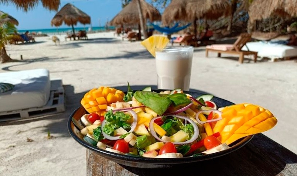 Una ensalada tropical con mango y una bebida refrescante se disfrutan en una idílica playa con palapas y el mar azul de fondo.