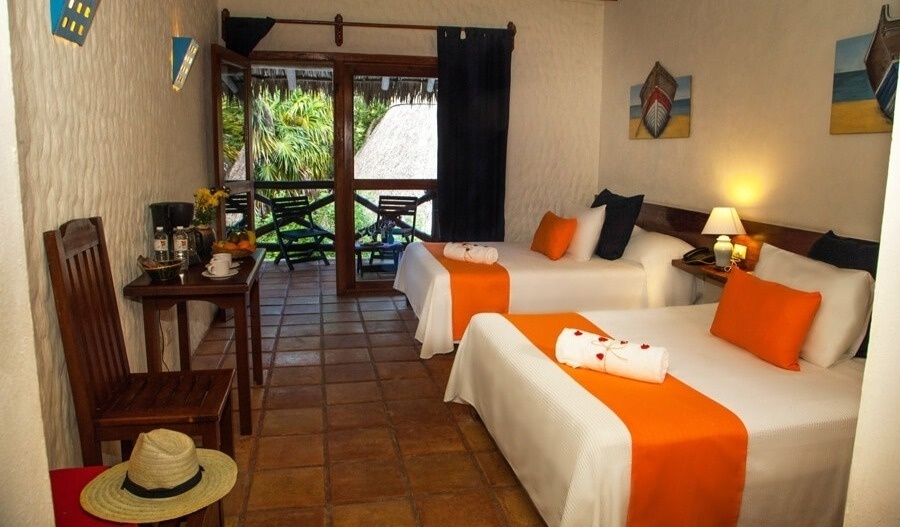 Una habitación de hotel de estilo rústico presenta dos camas con ropa de cama blanca y detalles naranjas, una mesa con cafetera y un balcón que da a una exuberante vegetación.