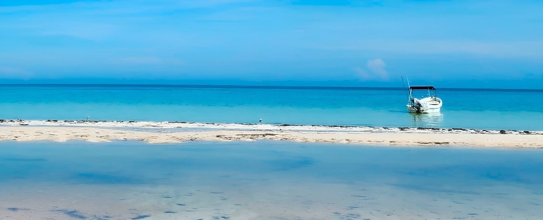 Una serena playa tropical presenta aguas cristalinas en tonos azules, una franja de arena blanca y un bote anclado en la distancia.