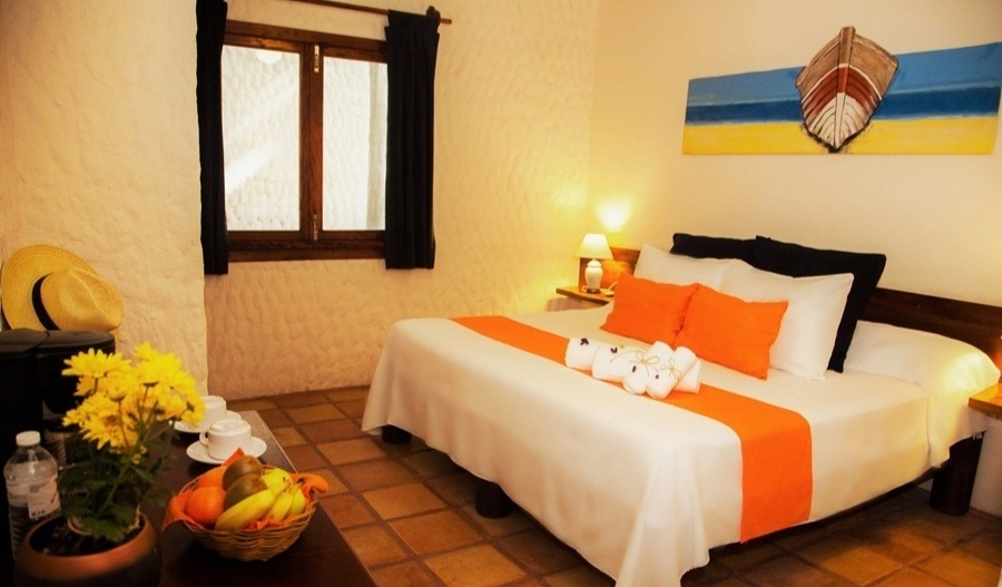Una acogedora habitación de hotel presenta una cama grande con sábanas blancas y detalles naranjas, una mesa con fruta y flores, y decoración de estilo costero.