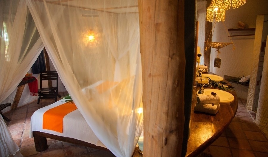 Un dormitorio rústico y acogedor presenta una cama con dosel mosquitera y un área de tocador doble con lavabos de cuenco en un ambiente cálido.