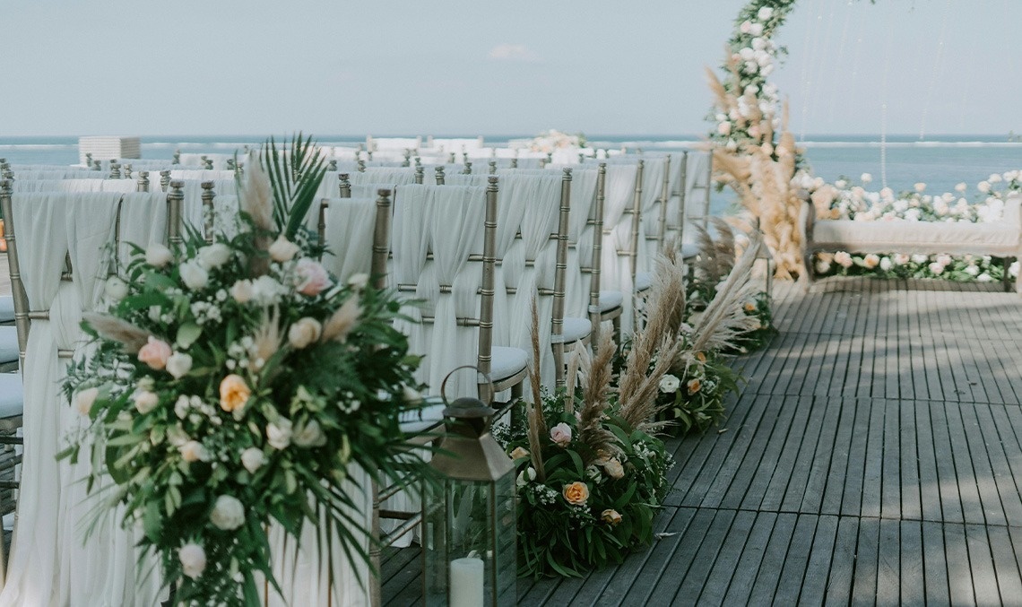 Elegante escenario de boda al aire libre junto al mar, con sillas cubiertas de blanco, arreglos florales con plumero y un camino de madera.