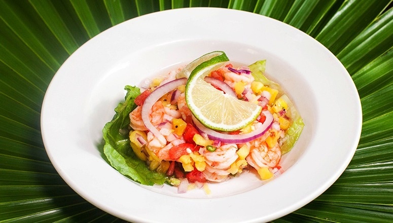 Un vibrante plato de ceviche tropical con camarones, mango, cebolla morada y lima se presenta en un tazón blanco sobre un fondo de hojas de palma verdes.