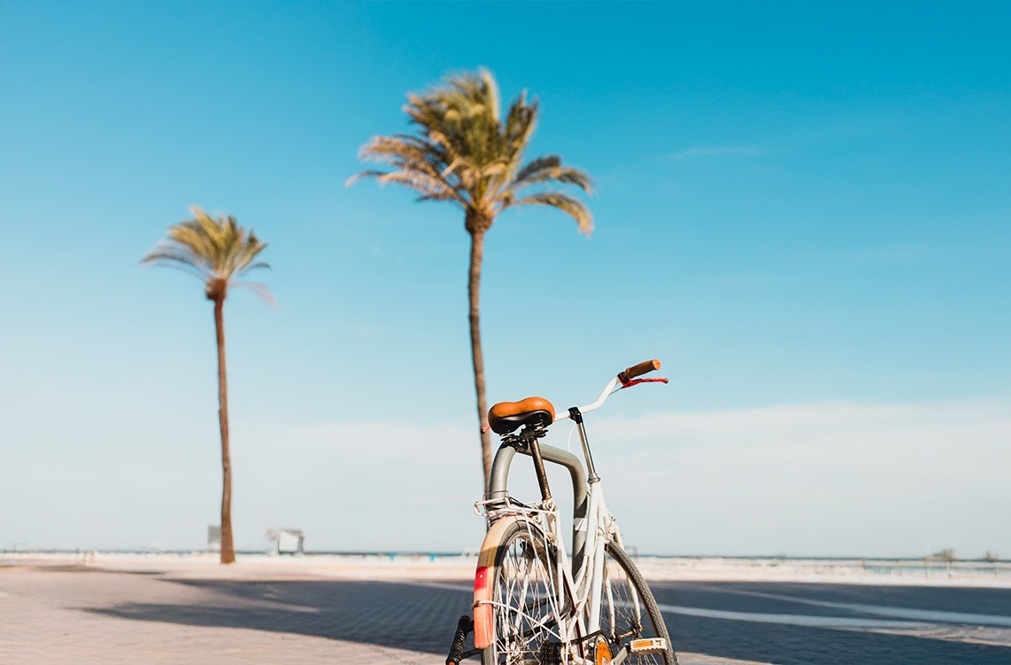 Una bicicleta blanca está aparcada en un paseo junto a palmeras bajo un cielo azul despejado.