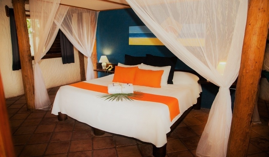 Un dormitorio de estilo tropical exhibe una cama con mosquitera, ropa de cama blanca acentuada con naranja y negro, paredes azules y un suelo de baldosas de terracota.
