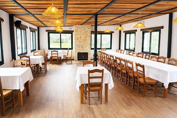 This image captures a rustic dining hall or event space featuring a long communal table and several smaller tables, all set with white tablecloths and wooden chairs, illuminated by woven pendant lights and natural light from multiple windows, with a stone fireplace and a central black pillar adding character to the room.