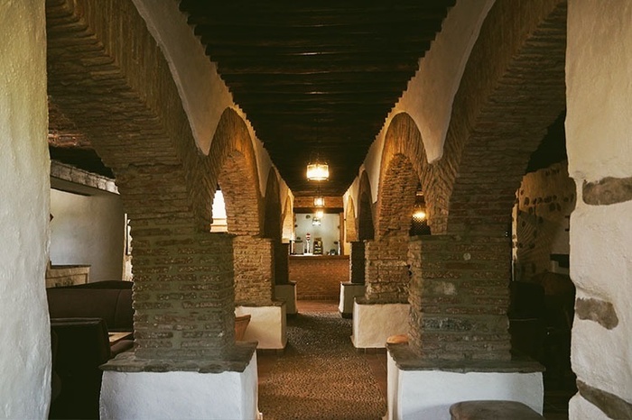 This image captures a rustic dining hall or event space featuring a long communal table and several smaller tables, all set with white tablecloths and wooden chairs, illuminated by woven pendant lights and natural light from multiple windows, with a stone fireplace and a central black pillar adding character to the room.