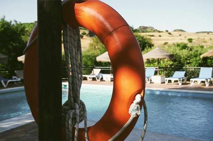 An orange lifebuoy with a coiled rope hangs prominently in the foreground, overlooking a sunny swimming pool lined with sun loungers and straw umbrellas, with natural hills visible in the background.