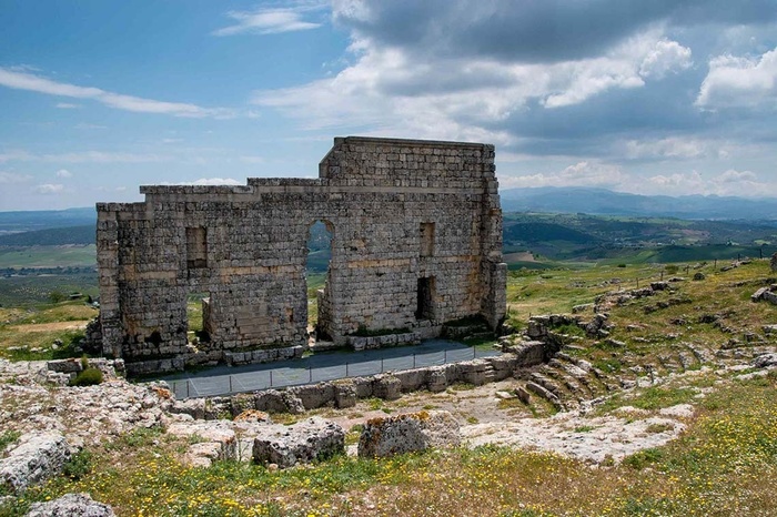 Antiguas ruinas de piedra con un teatro al aire libre, colinas verdes y cielo nublado. Un destino histórico.