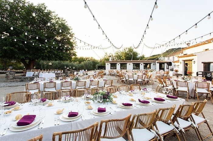 This image captures a rustic dining hall or event space featuring a long communal table and several smaller tables, all set with white tablecloths and wooden chairs, illuminated by woven pendant lights and natural light from multiple windows, with a stone fireplace and a central black pillar adding character to the room.