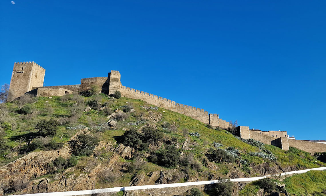 Castelo histórico em Horta de Bufenha, Portugal, com muralhas antigas numa colina verde sob céu azul.