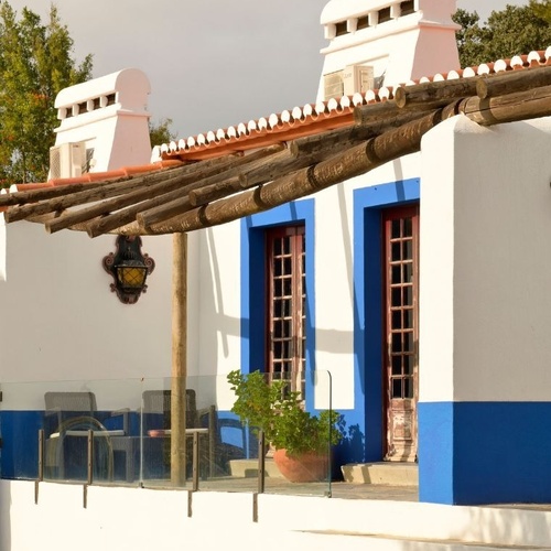 Uma casa tradicional branca e azul com telhado de telhas, uma pérgola de madeira e um terraço com escadas, iluminada pelo sol.