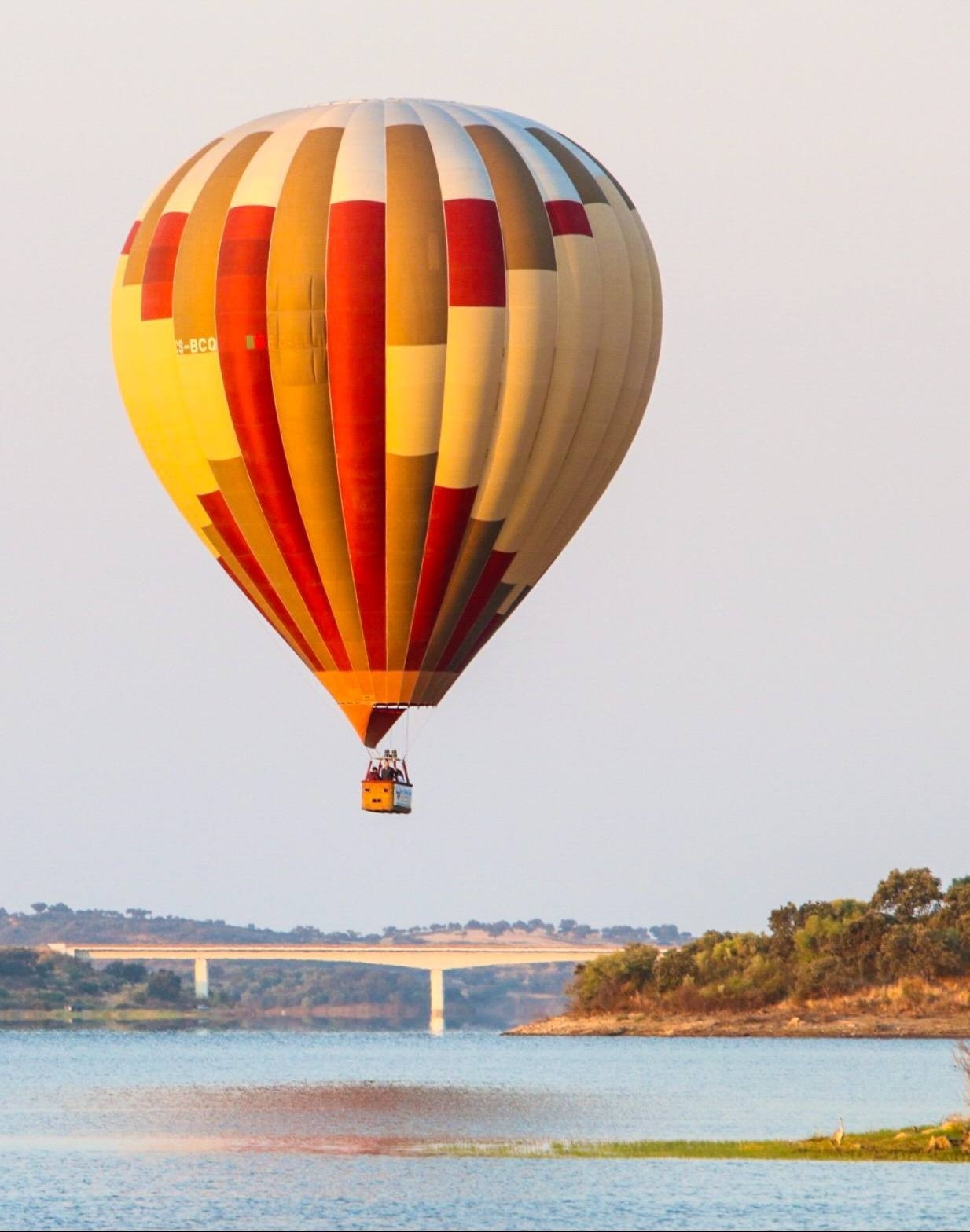 a hot air balloon flying over a body of water with a bridge in the background