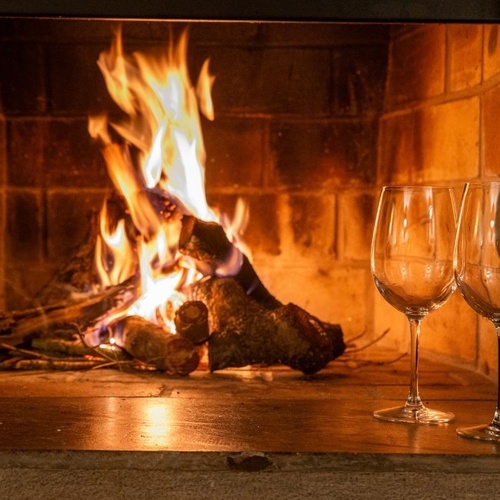 Uma lareira acesa com lenha em chamas, acompanhada por duas taças de vinho vazias e uma garrafa de vinho tinto, cria um ambiente acolhedor.