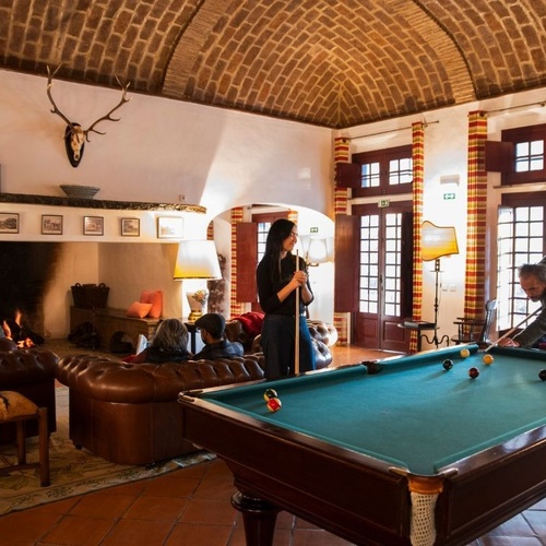 Num salão com teto de tijolos abobadado, pessoas jogam bilhar enquanto outras relaxam junto à lareira.