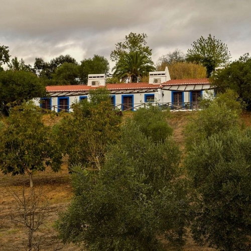 Uma casa branca com telhado de telha vermelha e detalhes em azul destaca-se entre a vegetação verdejante de uma encosta sob um céu nublado.