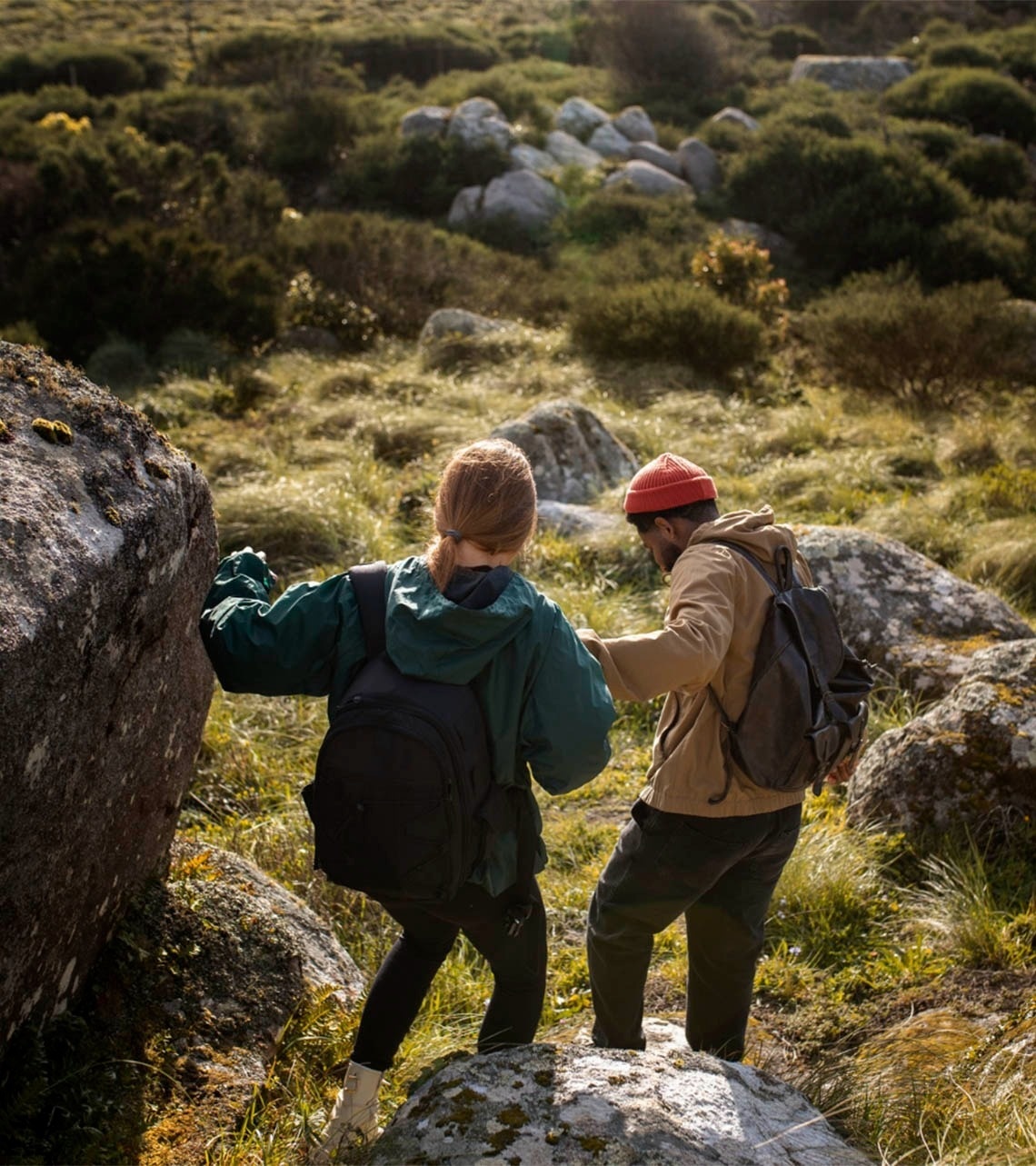 Dos personas, vistas de espaldas, caminan por un terreno rocoso y cubierto de hierba, equipadas con mochilas para una excursión.