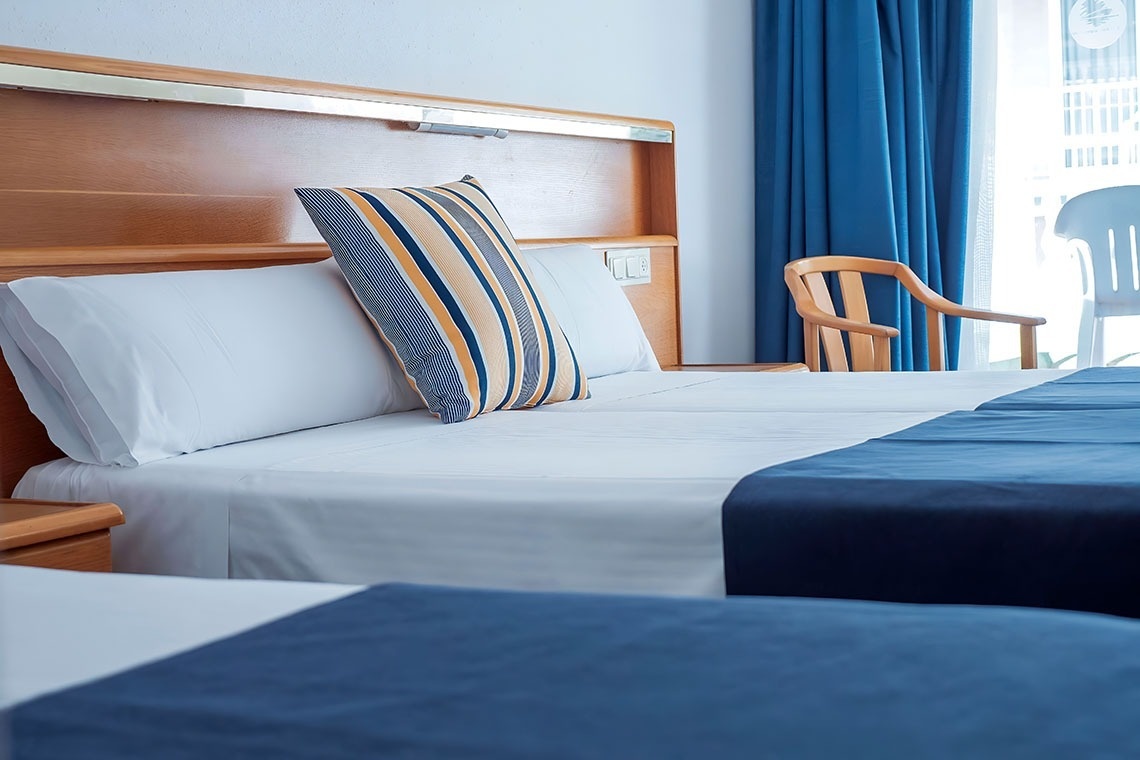 Une chambre d'hôtel lumineuse présente deux lits avec des draps blancs, des chemins de lit bleus, un oreiller à rayures colorées, une tête de lit en bois et des rideaux bleus près d'une fenêtre.