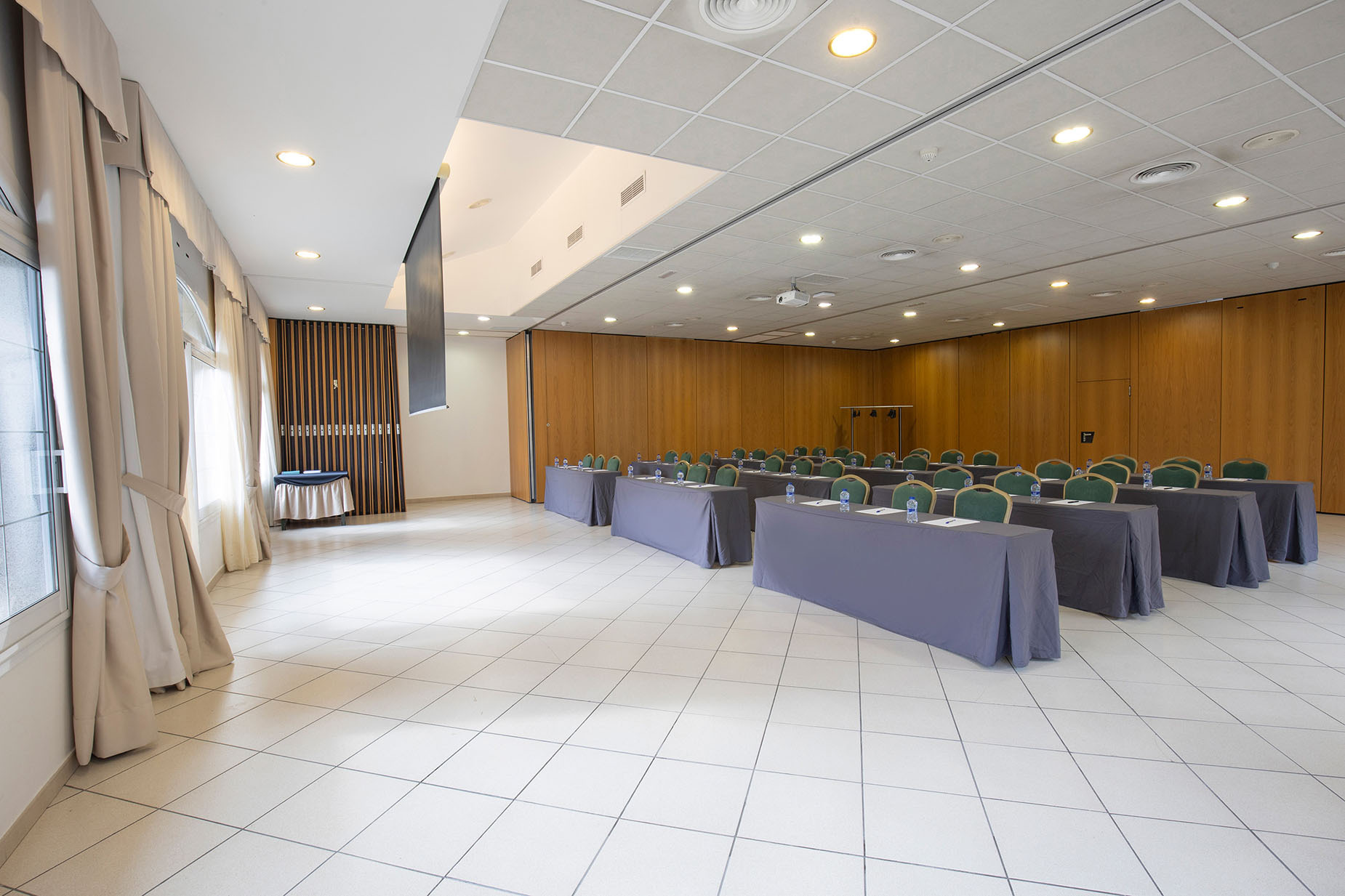 Salón de eventos preparado con mesas y sillas para una conferencia, con proyector y pantalla. Foto de NEXTA: Corporativa.