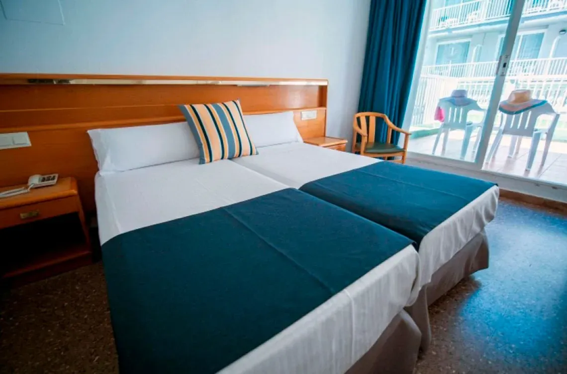 Une chambre d'hôtel lumineuse présente deux lits avec des draps blancs, des chemins de lit bleus, un oreiller à rayures colorées, une tête de lit en bois et des rideaux bleus près d'une fenêtre.