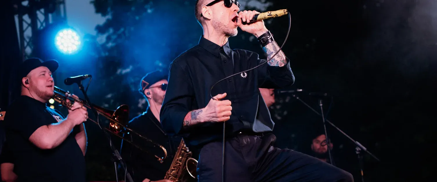 Un groupe joue sur scène sous des lumières bleues. Un chanteur porte une chemise noire et tient un micro. Un tromboniste et un saxophoniste sont à ses côtés. Activités | Nexta | Andorre | Site Officiel