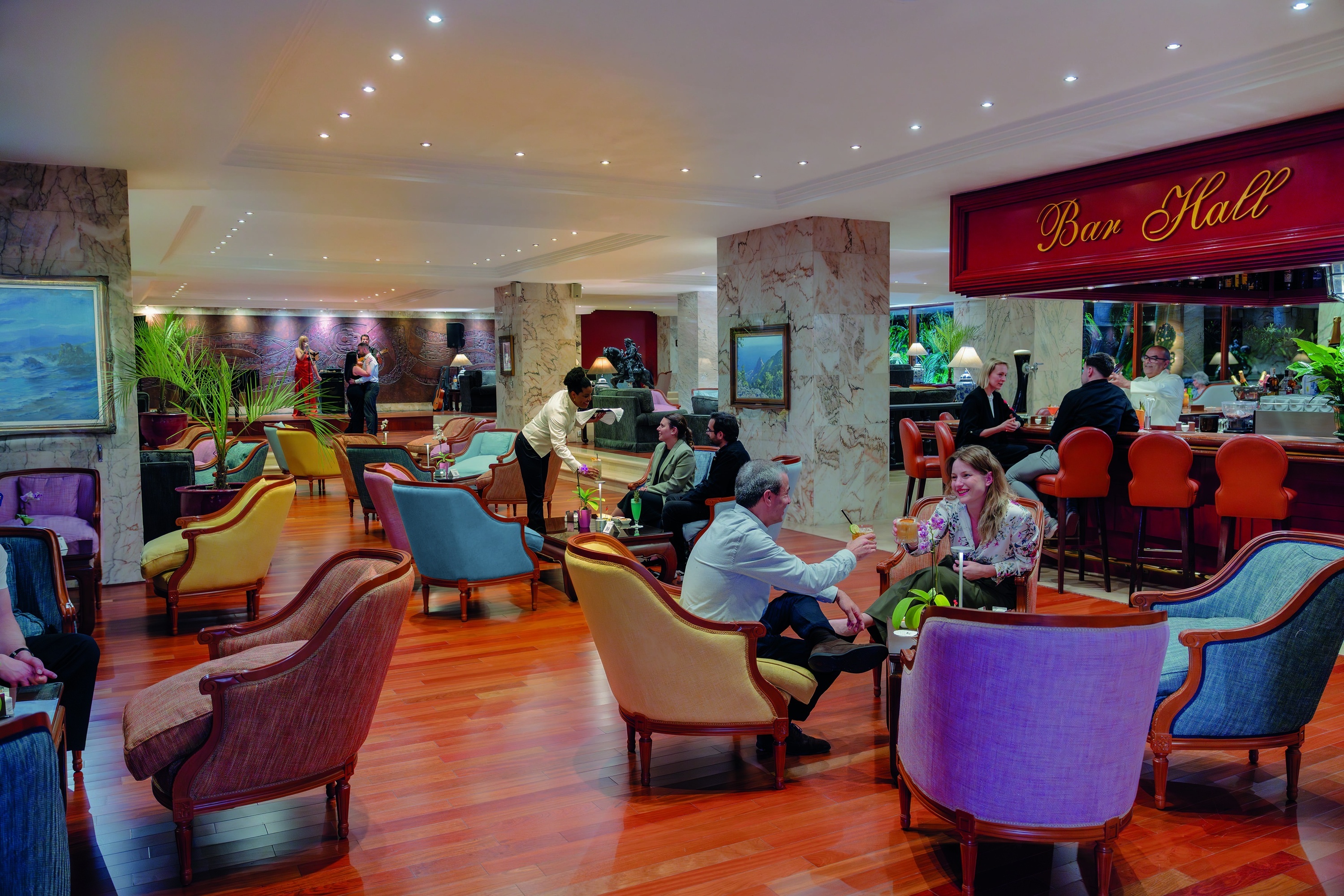 Un animado salón o bar cuenta con personas disfrutando de bebidas, un camarero sirviendo, una banda en vivo y un cartel que dice "Bar Hall".