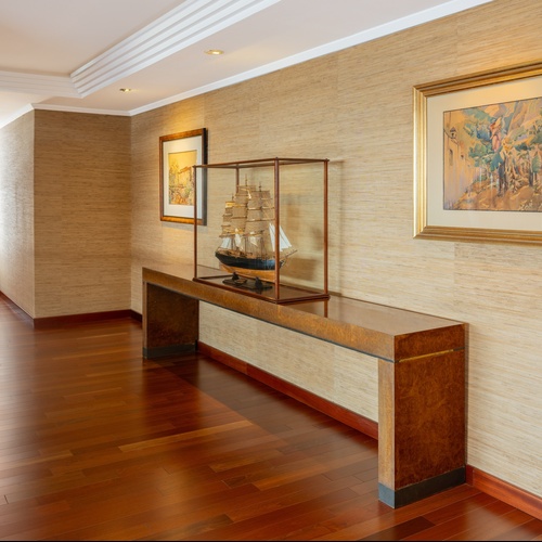 it: Un elegante corridoio illuminato con pavimento in legno, pareti rivestite in fibra naturale, una console con un modellino di veliero e dipinti, e una ringhiera curva.