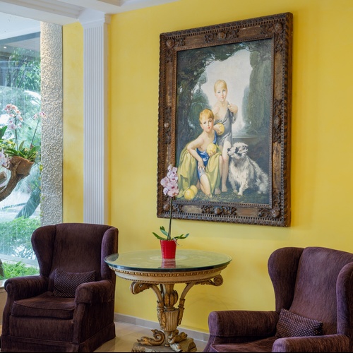 Un interno elegante presenta una parete gialla vivace con un grande dipinto classico, due poltrone marroni e un tavolino con un'orchidea, il tutto illuminato da un'ampia vetrata che mostra un lussureggiante giardino di orchidee e vegetazione.