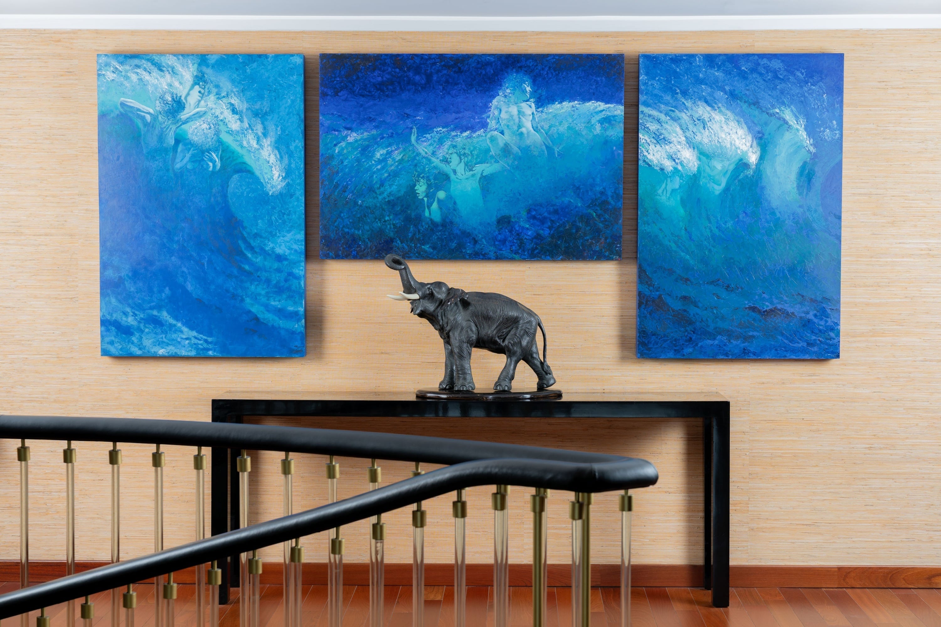 Tre blåa tavlor med vågor och figurer hänger över ett svart bord med en elefantskulptur, framför en ljus texturerad vägg och med ett trappräcke i förgrunden.