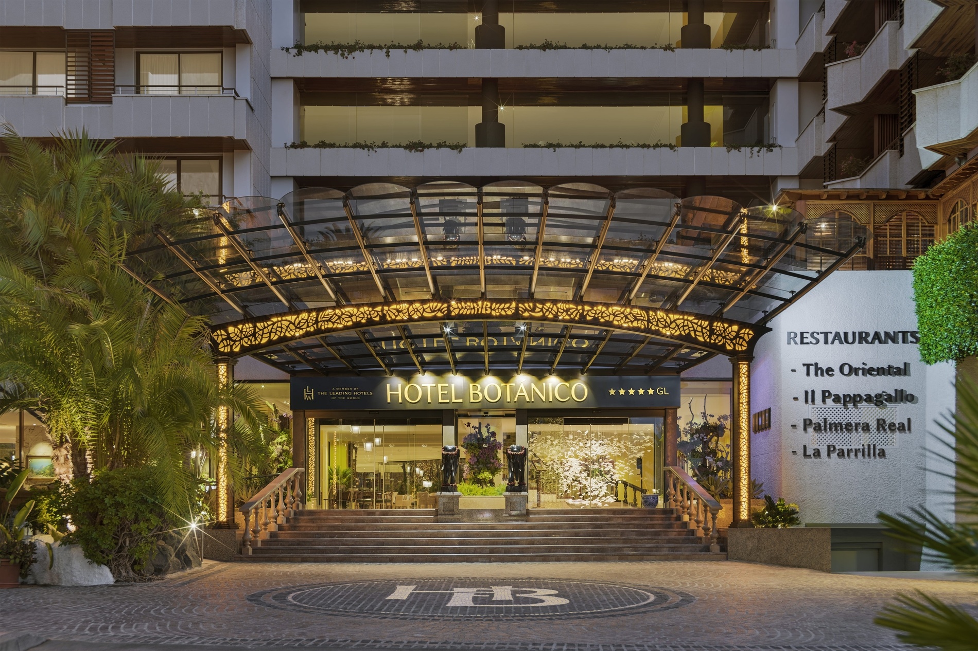 An aerial shot showcases a luxurious resort pool area with a curvilinear blue pool, surrounded by numerous lounge chairs, vibrant red umbrellas, and lush palm trees.