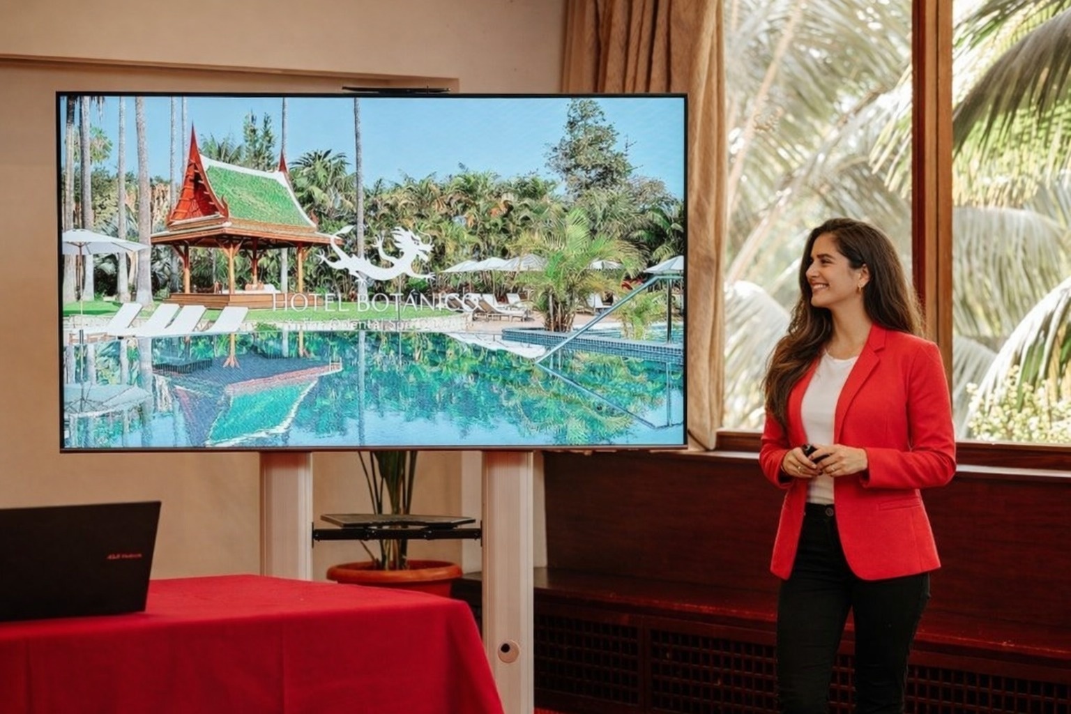 Een glimlachende vrouw in een zwart pak presenteert voor een groot scherm waarop een tropisch zwembadlandschap van 'Hotel Botánico' te zien is, met een laptop op een rode tafel op de voorgrond.