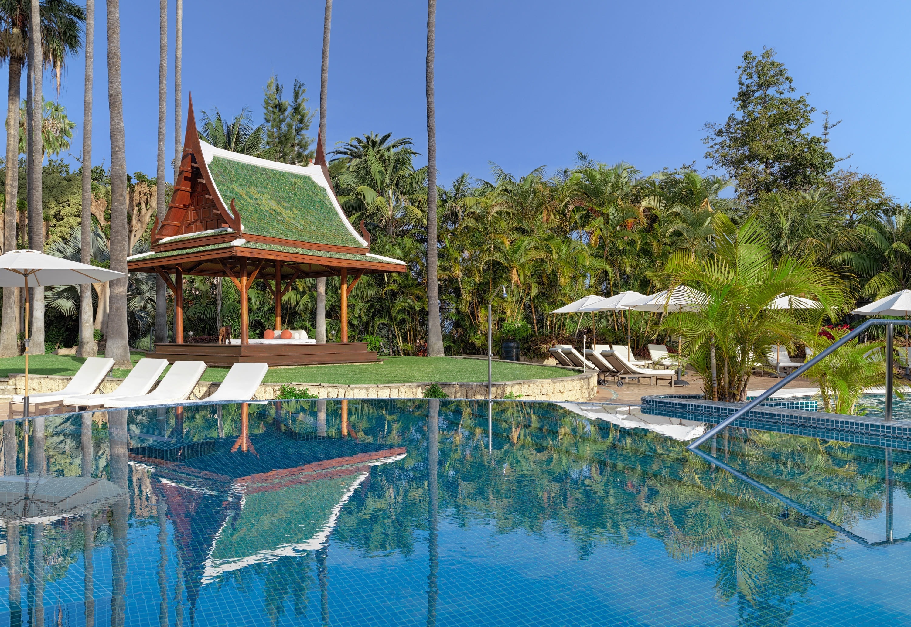 En thailändsk paviljong med grönt tak och palmer speglas i en blå kaklad pool bredvid solstolar under en klarblå himmel.
