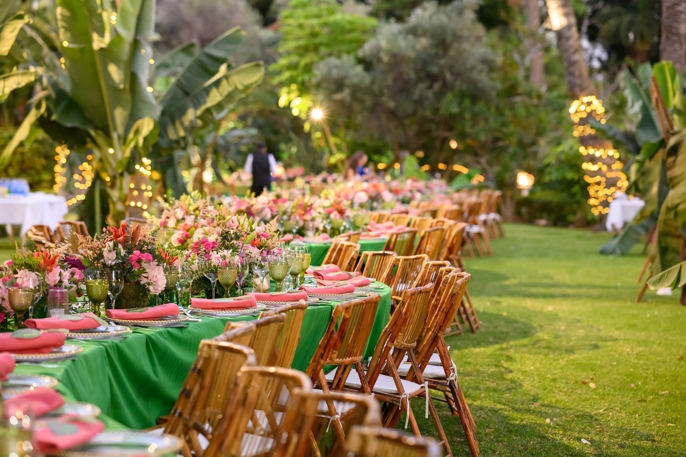 Ein festlich gedeckter Tisch mit Blumenarrangements und Holzstühlen steht in einem grünen Garten mit funkelnden Lichtern im Hintergrund.