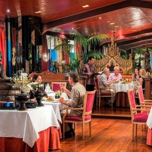 Guests are seen dining and being served by a waiter in a lavish, Asian-inspired restaurant featuring a rich red ceiling, ornate golden carvings, a Buddha statue, and lush greenery.