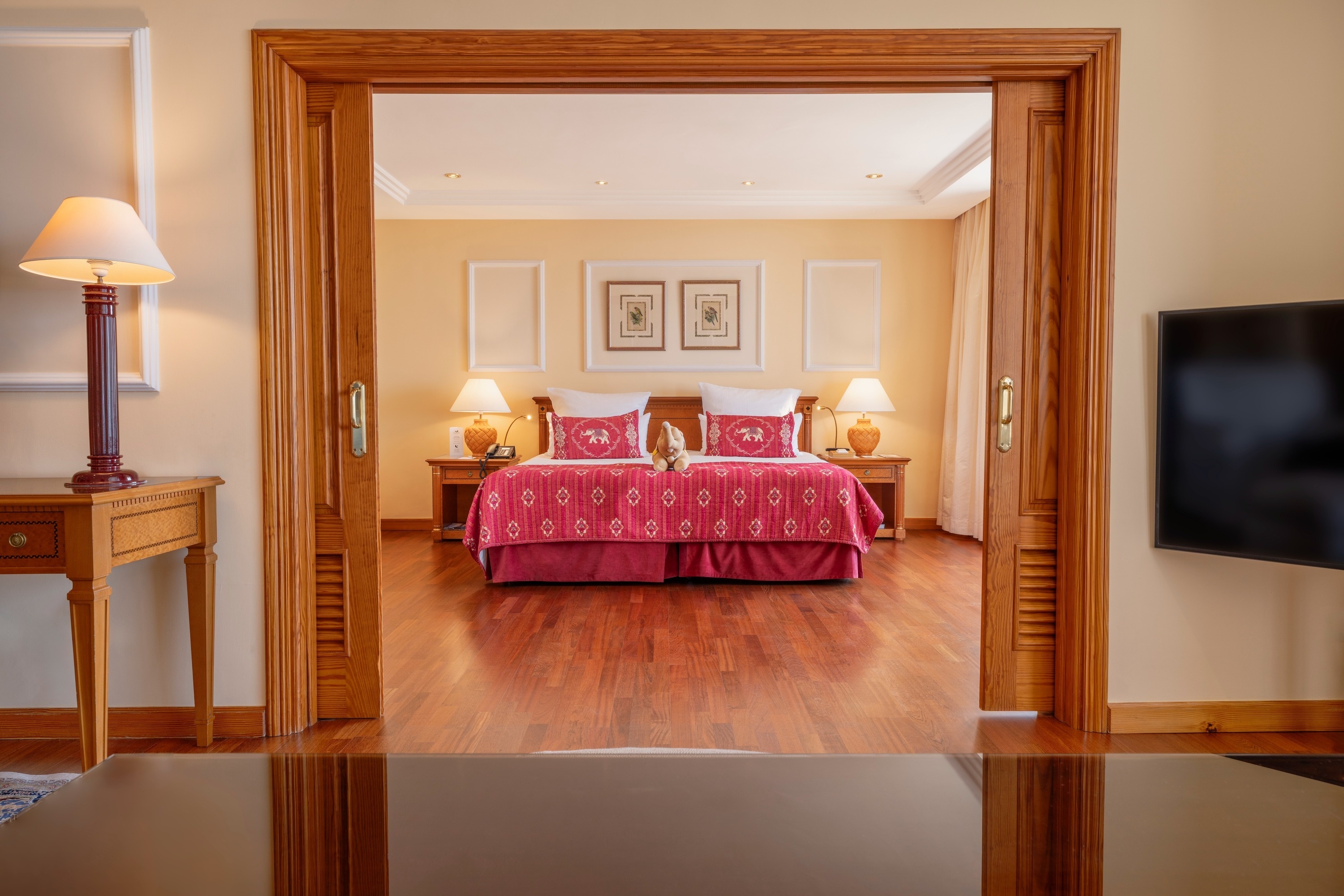 Ein offener, holzverkleideter Türrahmen führt den Blick in ein elegant eingerichtetes Schlafzimmer mit einem großen Bett, roten Tagesdecken und passenden Kissen.