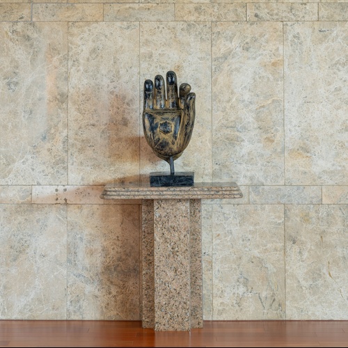 Una scultura scura a forma di mano posa su un tavolo di granito al centro di una parete piastrellata di marmo chiaro, con un pavimento in legno lucido in primo piano.