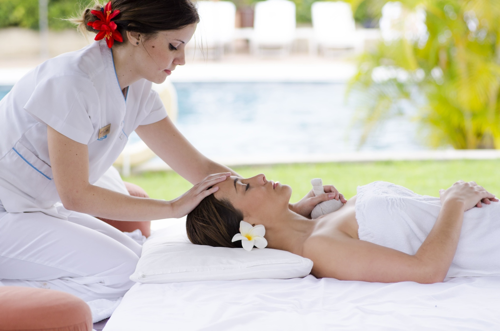 Un masseur donne un massage du dos à une personne, avec des mains posées sur la peau et des éléments de bien-être comme des pétales de roses rouges, un bol en bois et une petite fiole en premier plan.