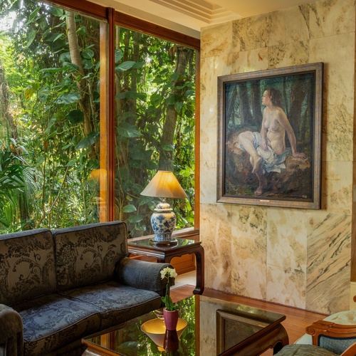 Una sala elegante presenta grandi finestre che si affacciano su una fitta vegetazione, un divano scuro e un quadro classico di figura femminile su una parete rivestita in marmo.