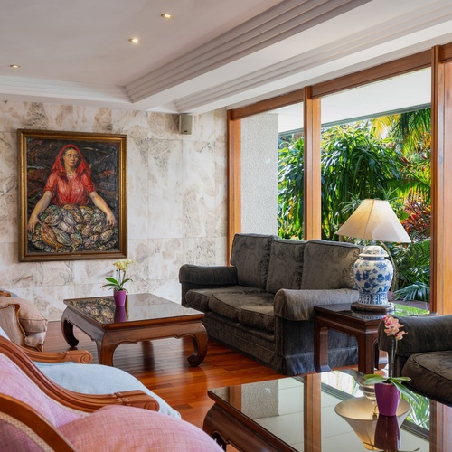 Un salotto elegante con pareti in marmo presenta un grande dipinto di una donna, mobili classici in legno e ampie finestre che si affacciano su una rigogliosa vegetazione.