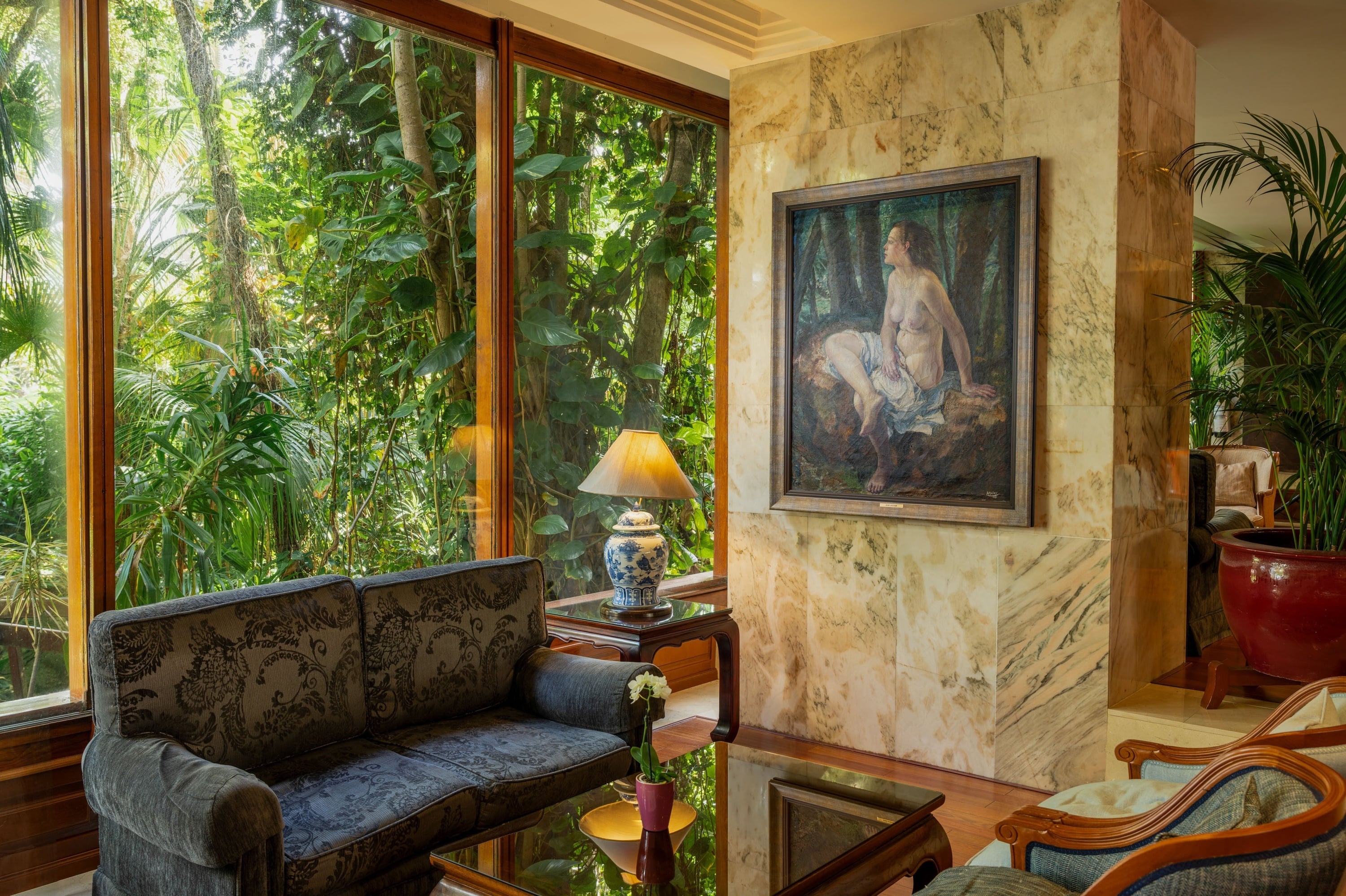 Ett elegant rum med stora fönster som vetter mot frodig tropisk grönska, möblerat med en soffa, en bordslampa och en målning på en marmorvägg.
