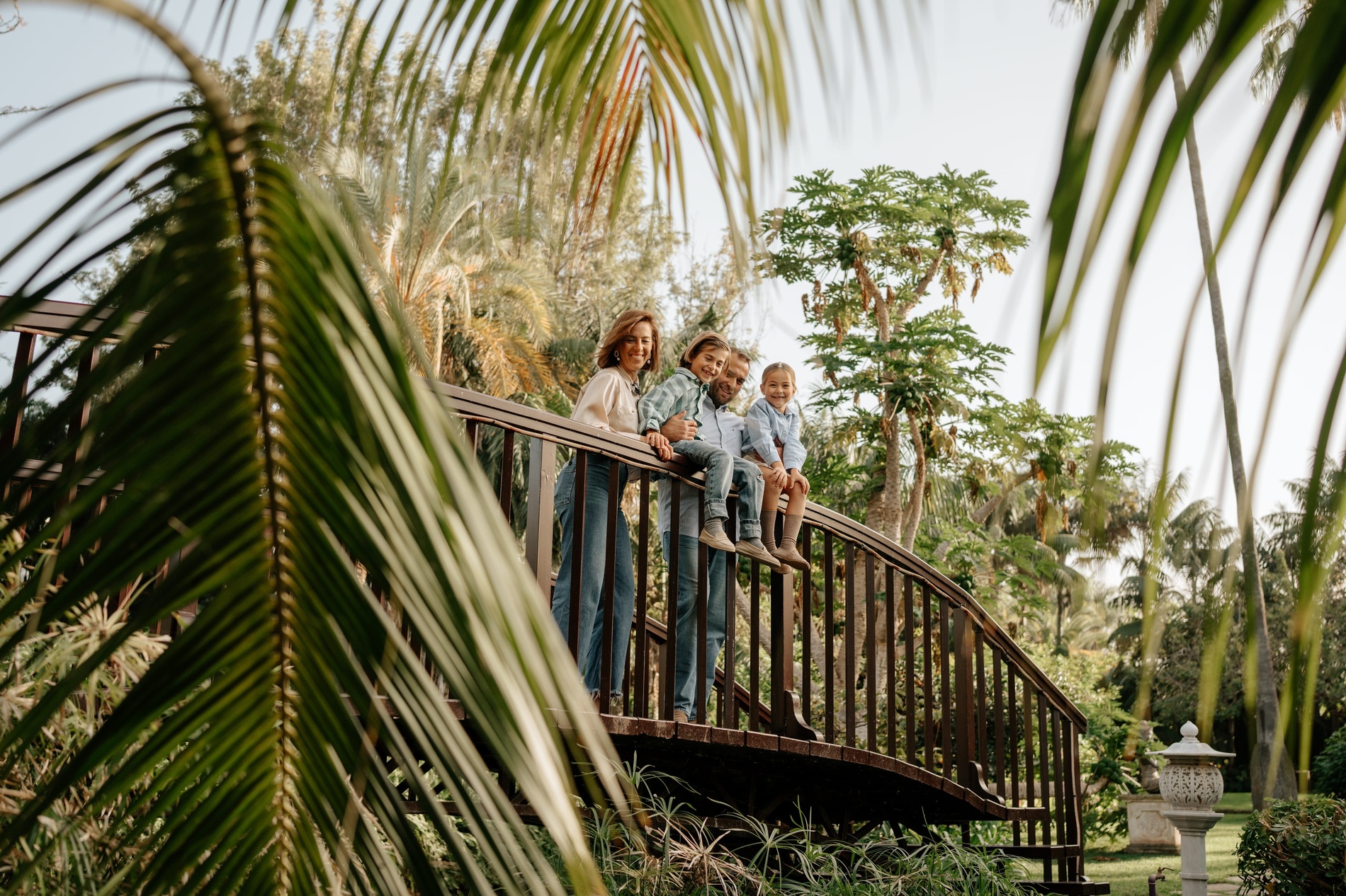 Una famiglia sorridente con due bambini che giocano allegramente nel prato, mentre i genitori li osservano da una panchina in un lussureggiante giardino tropicale con una fontana.