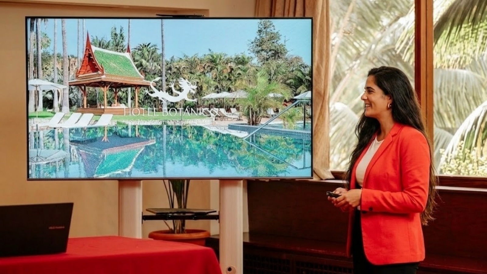 Une femme souriante, vêtue d'un blazer rouge, présente l'image d'un hôtel tropical ("Hotel Botanis") affichée sur un grand écran.