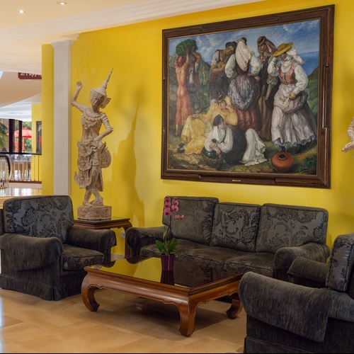 Una luminosa hall d'hotel presenta pareti gialle vivaci, un grande dipinto, due statue asiatiche e un'area salotto con un divano, poltrone e un tavolino in legno, con una scala a chiocciola visibile sullo sfondo.