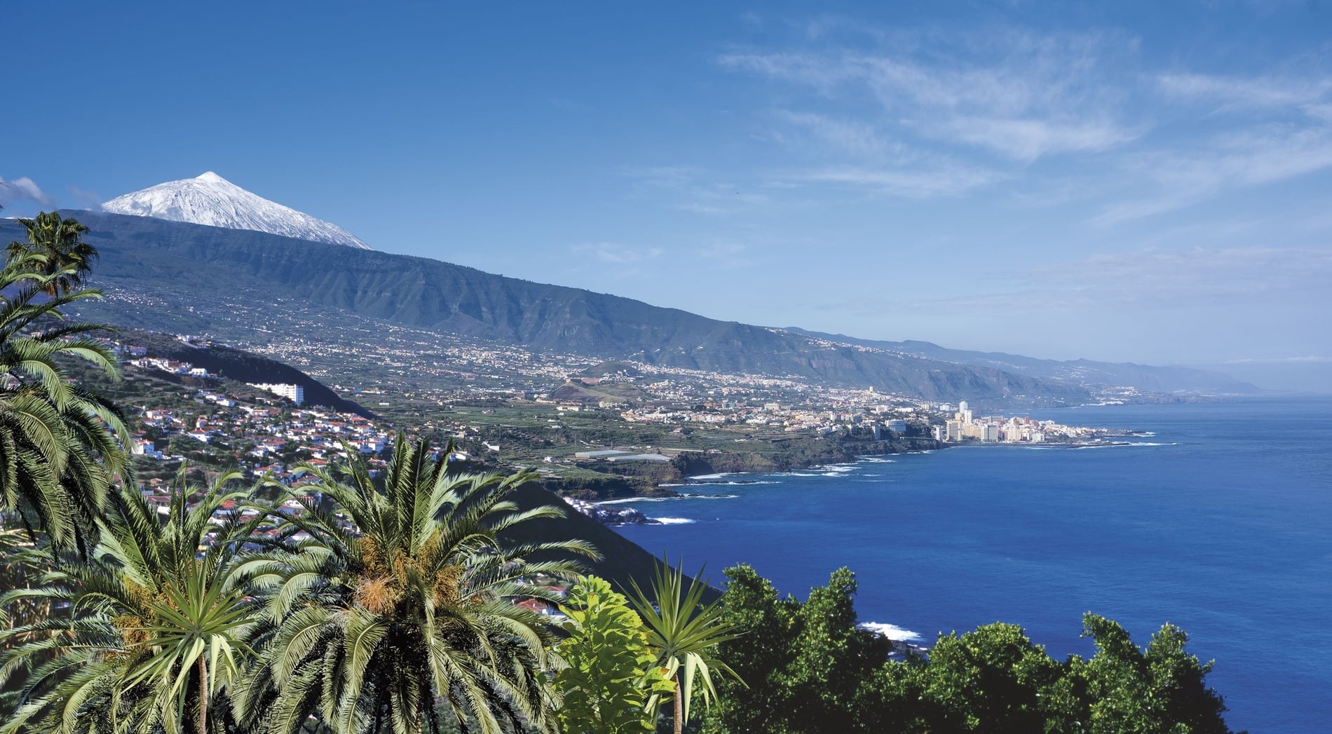 Ein sonniger Tag an einem schwarzen Sandstrand mit vielen Menschen und bunten Sonnenschirmen, im Hintergrund eine Küstenstadt und hohe Berge unter blauem Himmel.