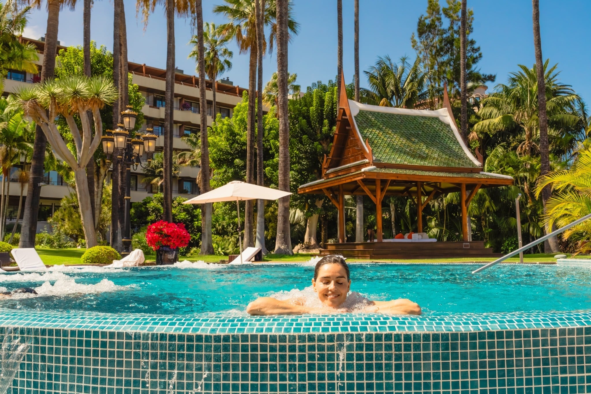 Una mujer sonríe relajada en un jacuzzi de azulejos azules en un complejo tropical con palmeras, un edificio hotelero y una cabaña de estilo asiático al fondo.