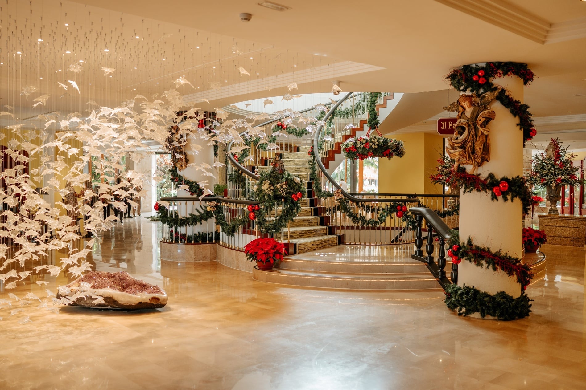 A grand, festive staircase decorated with lush green garlands, red ornaments, and bows ascends through an elegant interior, featuring a modern railing and transparent balusters.