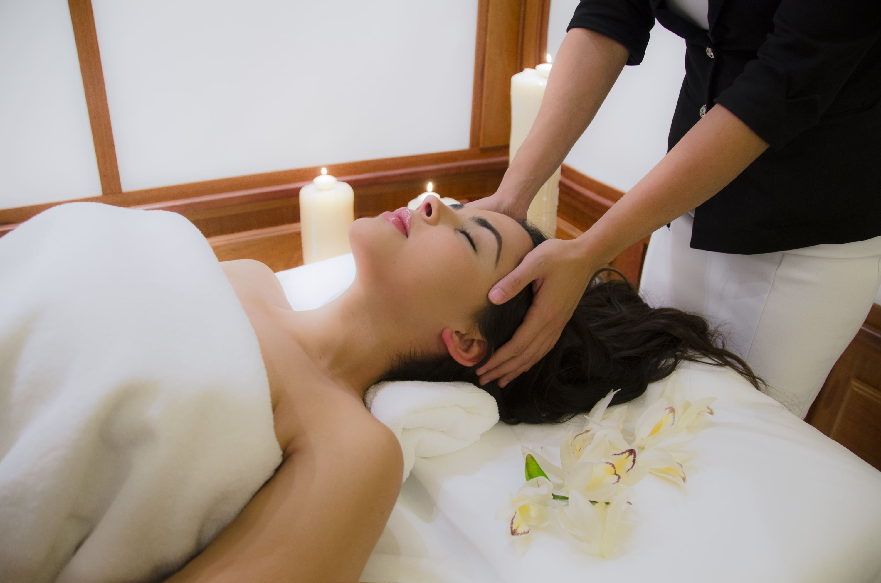 Una mujer yace relajada en una camilla de spa mientras recibe un masaje de cabeza, con velas encendidas y orquídeas blancas adornando el entorno.