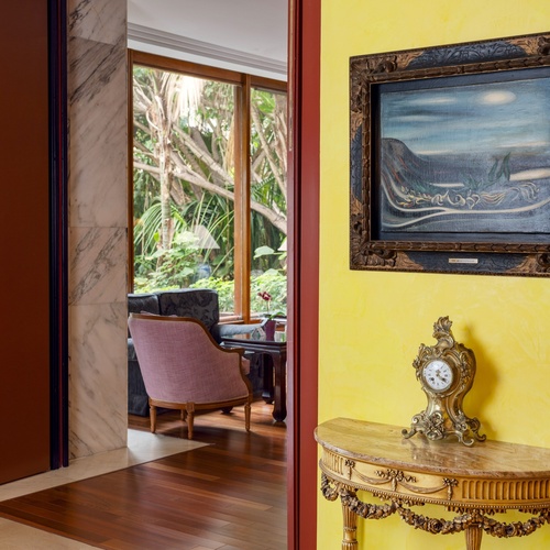 Un interno presenta un passaggio tra una parete gialla decorata con un quadro e una consolle con orologio, e un salotto con finestra che affaccia sulla natura e una poltrona rosa.
