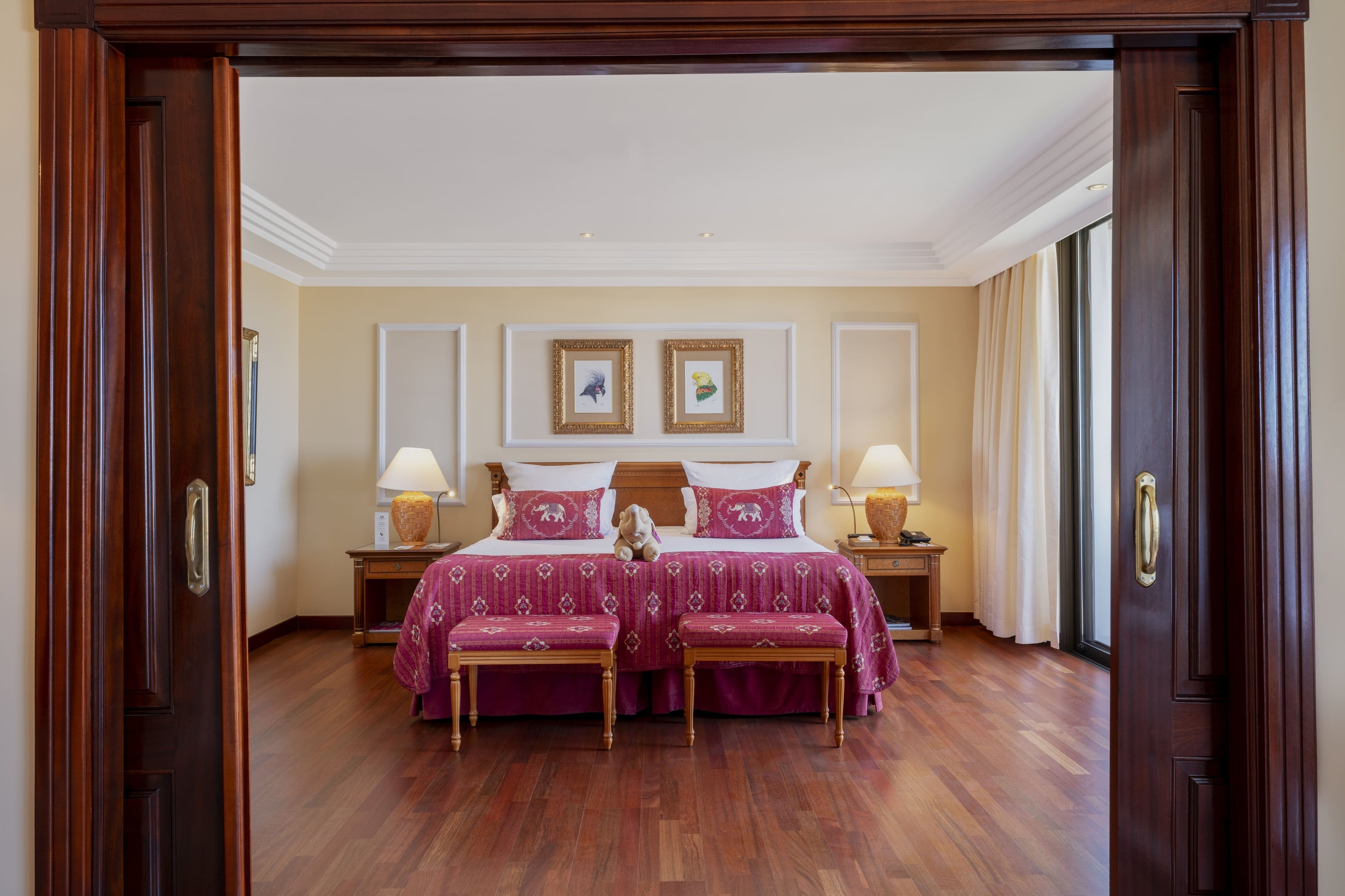 Ein elegant eingerichtetes Schlafzimmer mit einem Doppelbett und reichhaltigen roten Textilien, das durch eine offene dunkle Holztür gerahmt wird.