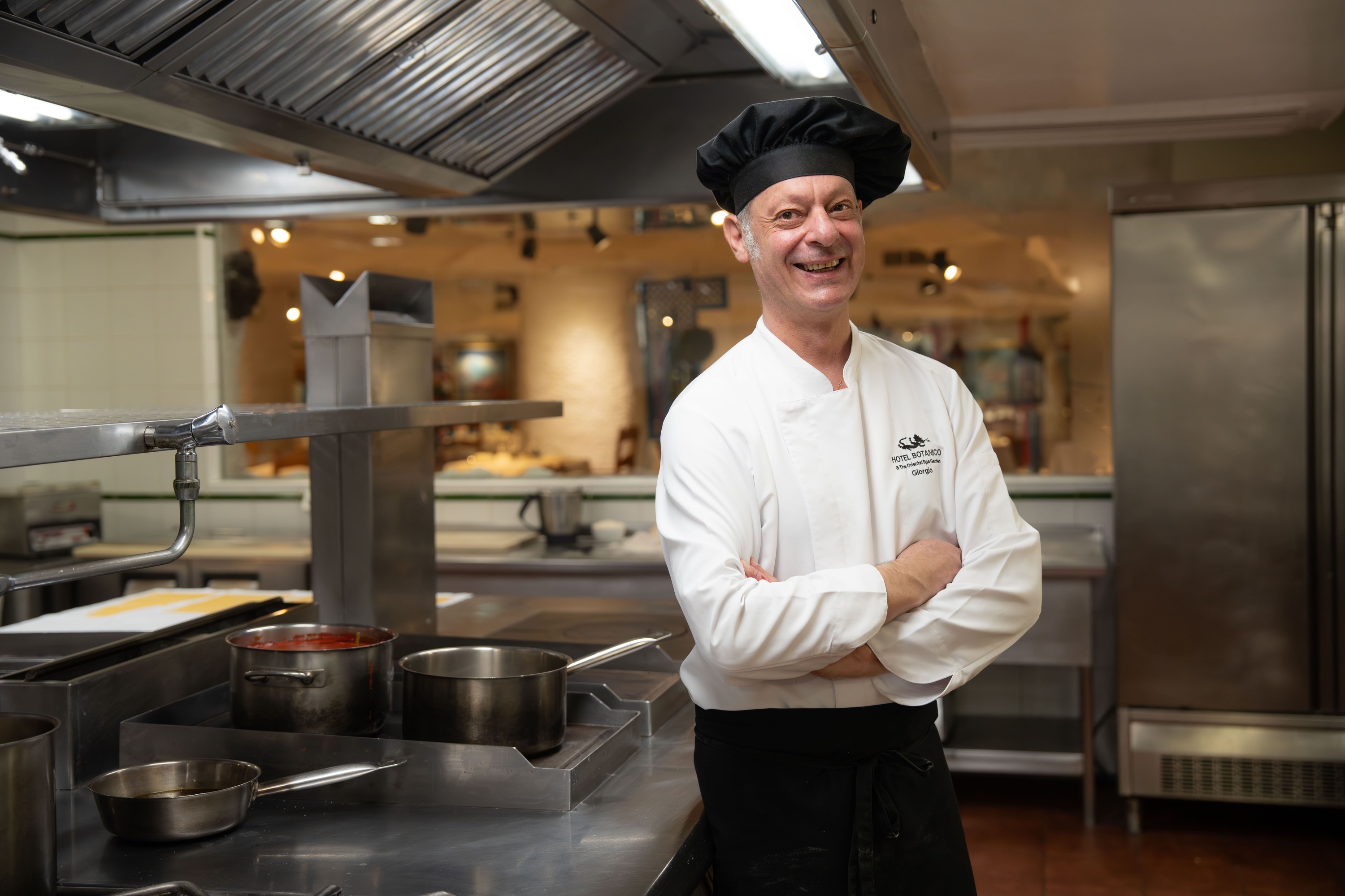 Een chef-kok in een wit uniform en zwarte muts plukt zorgvuldig groene kruiden uit een plantenbed in de buitenlucht.